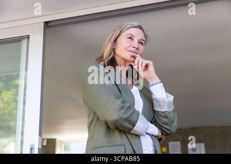 Femme senior portant blazer vert debout sur le patio, mur à côté de la porte vitrée Banque D'Images