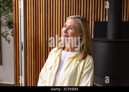 Femme senior debout à la maison par le mur à lattes à côté de la porte coulissante en verre avec poêle et plante Banque D'Images