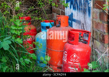Vieilles bouteilles de gaz rouges et bleues stockées à côté d'un mur de dépendance agricole, entourées de mauvaises herbes et de végétation envahie, Royaume-Uni Banque D'Images