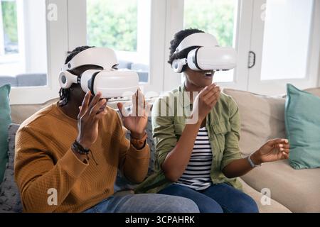 Couple afro-américain portant des casques vr soulevant et faisant des gestes sur le canapé dans le salon Banque D'Images