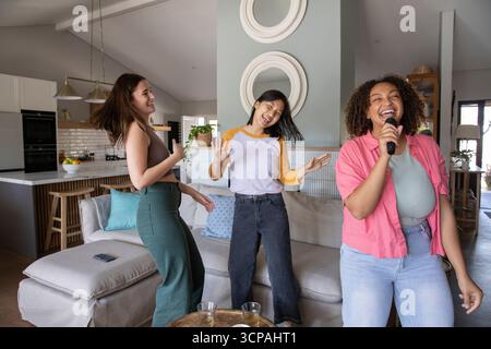 Diverses amies féminines chantant, dansant dans la cuisine du salon tenant le micro sans fil près du canapé Banque D'Images