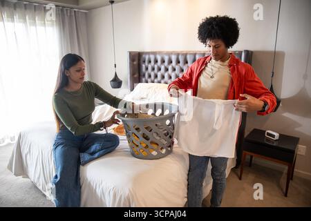 Couple diversifié tenant une chemise blanche et pliant le linge dans la chambre à coucher en utilisant le panier Banque D'Images