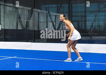 Athlète asiatique accroupie sur un court de padel bleu à la salle de gym tenant une raquette de padel noir, espace copie Banque D'Images