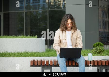 Jeune homme, étudiant, pigiste attrayant est assis sur le banc dans le parc urbain, en utilisant un ordinateur portable pour le travail à distance et l'éducation en ligne. À l'extérieur. Banque D'Images