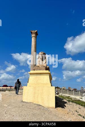 Alexandrie, Egypte - 13 février 2025 : statue de sphinx en granit avec pilier de Pompée à l'ancien Serapeum d'Alexandrie en Egypte sous un ciel bleu. Banque D'Images