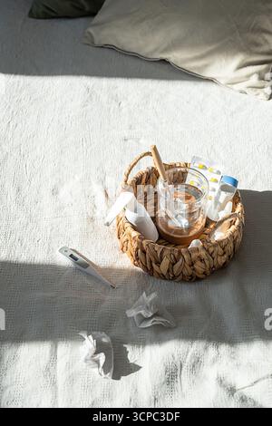 Panier de médicaments avec pilules, verre d'eau, thermomètre et mouchoirs usagés sur lit blanc. Concept de jour de maladie à domicile. Banque D'Images