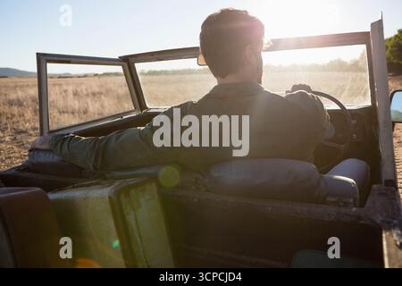 Homme adulte moyen conduisant une jeep saisissant le volant le long du chemin de terre à travers l'herbe à l'heure dorée Banque D'Images