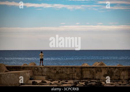 Une personne se tient sur une jetée en béton, tenant un téléphone ou un appareil photo, et regardant le vaste océan sous un ciel nuageux, capturant peut-être la mer sereine Banque D'Images