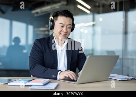 Jeune homme d'affaires asiatique assis dans le bureau portant des écouteurs et souriant tout en travaillant sur un ordinateur portable. Banque D'Images