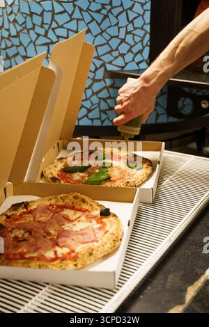 On voit une main couler de l'huile d'olive sur une pizza Margherita avec des feuilles de basilic frais, tandis qu'une autre pizza prosciutto repose à côté dans une boîte en carton ouverte Banque D'Images