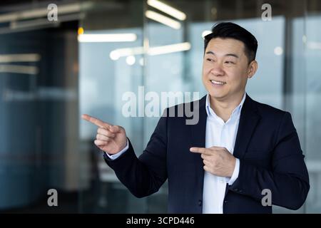 Asiatique souriant jeune homme d'affaires debout dans le bureau portant costume et pointant avec les doigts sur le côté à l'espace vide, la présentation et la publicité. Banque D'Images