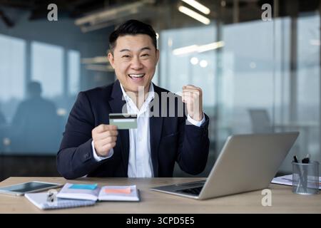 Portrait de jeune homme asiatique heureux en costume travaillant dans le bureau à l'ordinateur portable, tenant la carte de crédit, montrant le geste de victoire et regardant la caméra. Banque D'Images