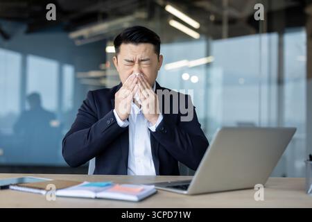 Malade jeune homme asiatique est assis au bureau devant un ordinateur portable dans le bureau dans un costume d'affaires et s'essuie le nez, avec une serviette d'un nez qui coule. Banque D'Images