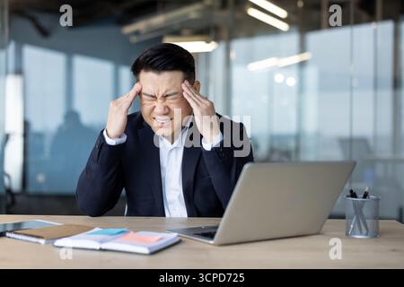 Un jeune homme asiatique est assis à son bureau et grimace dans la douleur, tenant sa tête dans ses mains. Banque D'Images