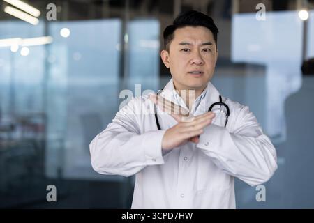 Portrait d'un jeune médecin asiatique sérieux et inquiet dans un manteau blanc debout dans le hall de la clinique et montrant un panneau d'arrêt avec ses mains à la caméra. Banque D'Images