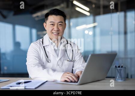 Portrait d'un jeune médecin professionnel asiatique souriant travaillant dans un hôpital, assis dans le bureau à une table avec un ordinateur portable et regardant la caméra. Banque D'Images