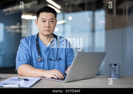 Portrait d'un jeune médecin asiatique assis dans un bureau de clinique, travaillant sur un ordinateur portable et regardant sérieusement la caméra. Banque D'Images