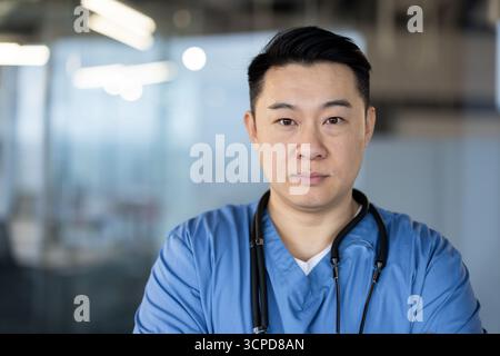 Portrait en gros plan d'un jeune médecin asiatique, infirmier debout dans un hôpital en uniforme bleu et avec un stéthoscope, regardant sérieusement la caméra. Banque D'Images