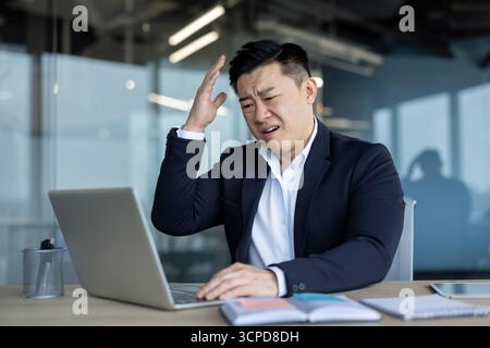 Frustré et contrarié jeune homme asiatique assis dans le bureau au bureau et travaillant sur ordinateur portable faisant des gestes avec la main. Banque D'Images