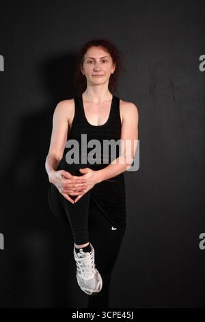 Jeune femme en vêtements de sport noirs étirant la jambe en studio, entraînement de fitness, style de vie sain et concept d'exercice sur fond sombre Banque D'Images