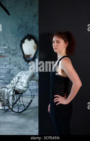 Jeune femme en sportswear noir debout latéralement à l'intérieur, portrait de fitness et de style de vie avec mur sombre et fond intérieur vintage Banque D'Images