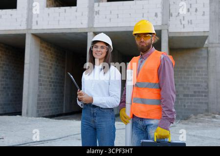 Architecte et contremaître en casques durs sourient à un chantier de construction, montrant le travail d'équipe et la sécurité. Les plans et le presse-papiers soulignent le souci du détail Banque D'Images