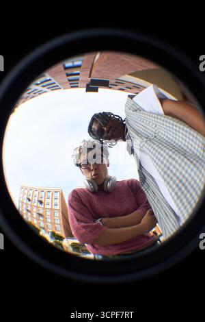 Vue de poisson de deux jeunes hommes, caucasiens avec des cheveux bouclés et des lunettes, noir avec des cheveux tressés, debout à l'extérieur dans un cadre urbain, regardant vers le bas dans la caméra avec des expressions sérieuses Banque D'Images