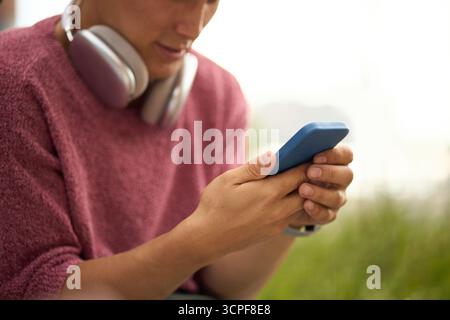 Jeune homme caucasien tenant un smartphone avec les deux mains, portant des écouteurs autour du cou, assis à l'extérieur, se concentrant sur l'écran de l'appareil, visage partiel visible Banque D'Images