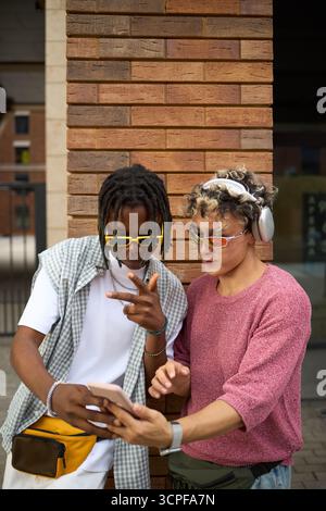 Deux jeunes hommes, des amis noirs et caucasiens, debout dehors en utilisant un smartphone ensemble, portant des lunettes de soleil et des écouteurs, interagissant et regardant l'écran de l'appareil Banque D'Images