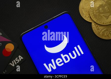 Dhaka, Bangladesh- 26 août 2025 : smartphone affichant le logo webull entouré de pièces Bitcoin et de cartes de paiement Banque D'Images