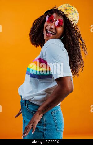 Jeune femme latine insouciante portant un t-shirt arc-en-ciel, un chapeau drôle et des lunettes roses pose avec un fond orange, elle a les cheveux bouclés et sourit Banque D'Images