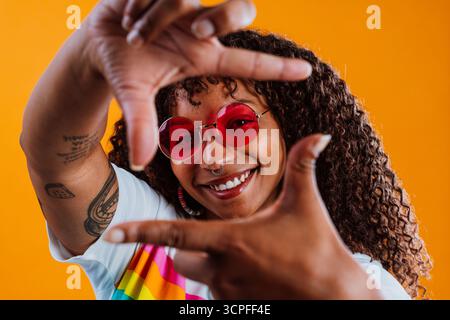 Jeune artiste souriante avec les cheveux bouclés et piercing nez portant des lunettes de soleil rouges et un t-shirt lgbtq+ encadrant son visage avec les mains tout en posant sur BA orange Banque D'Images