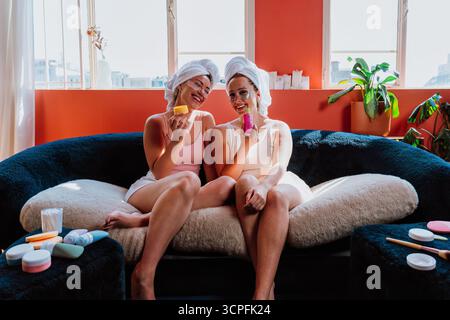 Deux femmes joyeuses avec des serviettes sur la tête et des patchs sous les yeux sont assises sur un canapé confortable, riant et tenant des produits de beauté, appréciant Banque D'Images