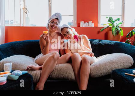 Deux femmes joyeuses avec des serviettes sur la tête et des patchs sous les yeux sont assises sur un canapé confortable, riant et tenant des produits de beauté, appréciant Banque D'Images