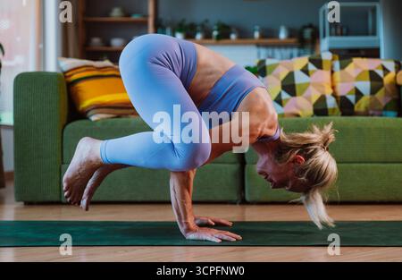 Femme senior dans Activewear maintient l'équilibre dans la posture du corbeau, démontrant la force et la flexibilité pendant une séance de yoga à la maison sur un tapis vert Banque D'Images