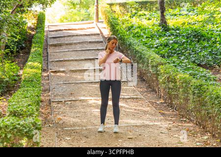 Coureur se reposant après l'exercice dans un parc luxuriant, prenant un moment pour vérifier la fréquence cardiaque sur sa smartwatch, en se concentrant sur la santé, le bien-être et les objectifs de fitness d Banque D'Images