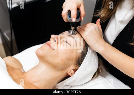 La cliente aime le traitement de rajeunissement facial de levage RF, couché confortablement sur le lit de traitement, tandis que l'esthéticienne applique le dispositif pour l'improvem de peau Banque D'Images