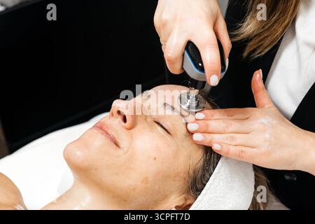 La cliente profite du traitement de rajeunissement facial de levage RF, couché confortablement sur le lit de traitement, tandis que l'esthéticienne applique le dispositif pour le resserrement de la peau Banque D'Images
