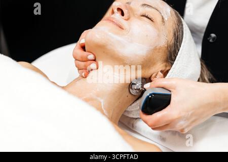 La cliente profite du traitement de rajeunissement facial de levage RF, allongée sur une table de traitement, avec une atmosphère apaisante et des mains professionnelles fournissant CA Banque D'Images