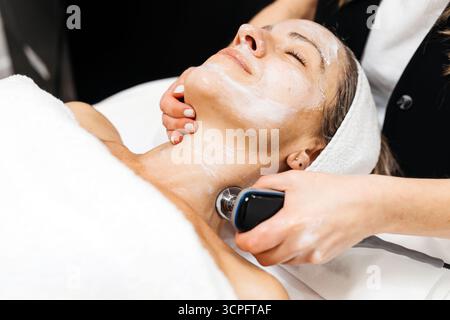 La cliente profite du traitement de rajeunissement facial RF Lifting, allongée sur une table de traitement, entourée d'une atmosphère apaisante et d'un outil de soins de la peau avancé Banque D'Images