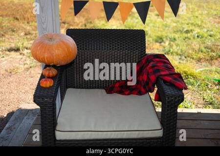 Patio extérieur décoré pour Halloween. Une chaise en osier se trouve sur un porche avec une couverture, des citrouilles et une bannière d'Halloween en arrière-plan. Banque D'Images