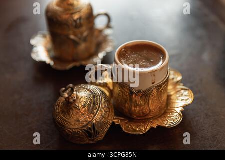 Café turc. Une tasse à café dorée luxueuse repose sur une soucoupe en filigrane assortie, avec une théière décorative subtilement visible en arrière-plan Banque D'Images