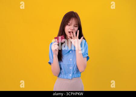Une jeune femme dans une blouse bleue tient une carte de crédit, ayant l'air surprise. Idéal pour des sujets comme le shopping, la finance, l'achat en ligne Banque D'Images