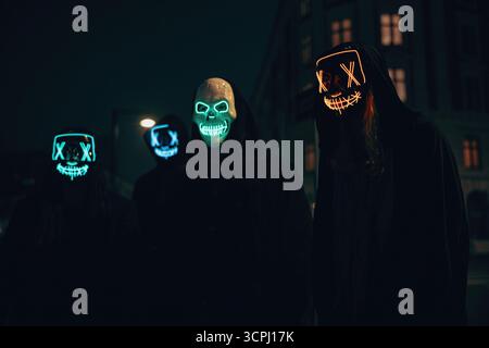 Groupe d'individus portant des masques lumineux la nuit dans un environnement urbain de rue, vêtus de pseudonymes. Banque D'Images