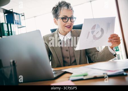 Femme d'affaires mature examine les documents de données d'entreprise dans un espace de bureau moderne, en analysant et en élaborant des stratégies de mesure de l'entreprise pendant le travail. Banque D'Images