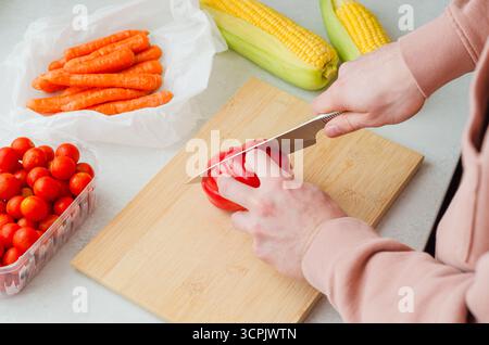 Gros plan d'un homme tranchant des tomates beefsteak côtelées sur une planche à découper en bois avec des légumes de ferme biologiques - maïs, carottes et tomates cerises. Banque D'Images