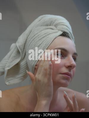 Une personne applique de la crème sur son visage dans une salle de bain contemporaine après une douche rafraîchissante, enveloppée dans une serviette. Banque D'Images