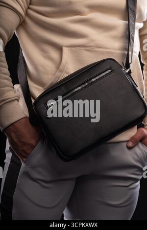 homme vêtu d'un sweat à capuche beige présente un sac noir soulignant son style décontracté. Il se tient devant un fond sombre ajoutant de la profondeur à la scène. Banque D'Images