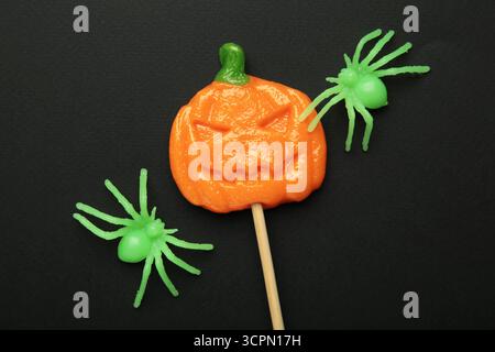 Sucettes d'Halloween en forme de citrouille avec araignée décorative sur fond noir. Gâteries festives pour les fêtes de fin d'année. Vue de dessus Banque D'Images