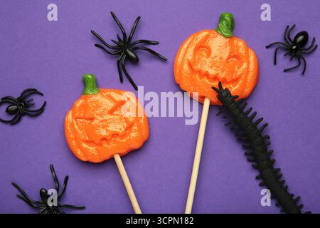 Sucettes d'Halloween en forme de citrouille avec araignée décorative sur fond violet. Gâteries festives pour les fêtes de fin d'année. Vue de dessus Banque D'Images